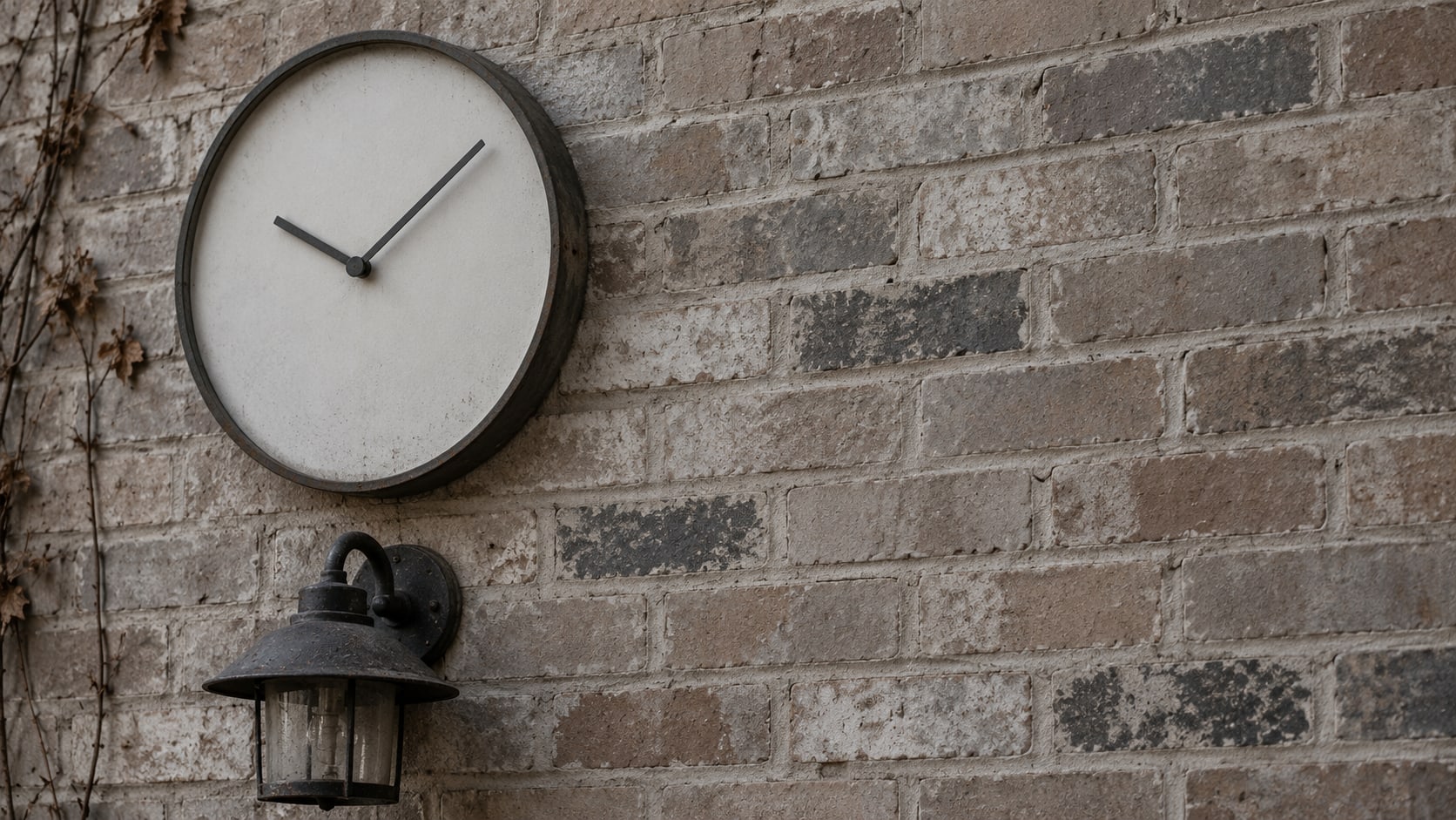 A plain wall clock without numbers hanging above an unlit porch lamp on a brick wall.