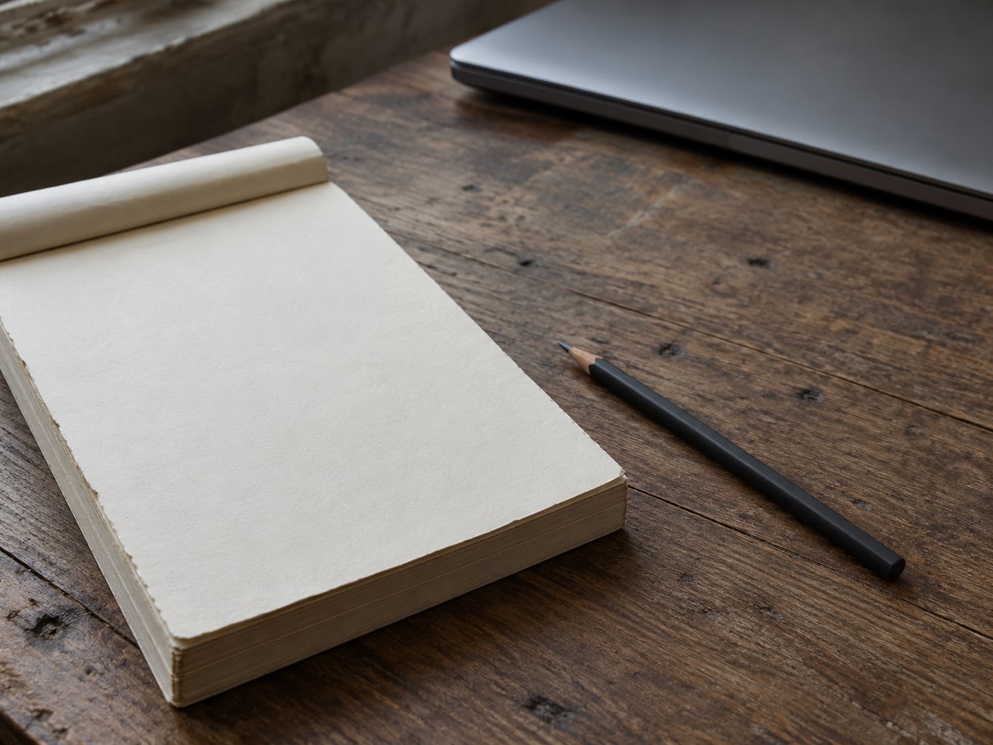 A blank notepad with a pencil beside a closed laptop on a wooden desk.