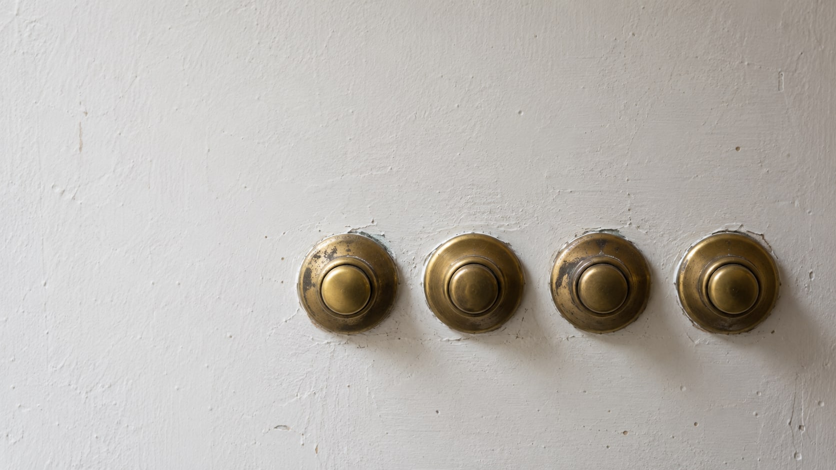 A row of four plain brass push buttons mounted on a bare painted wall panel.