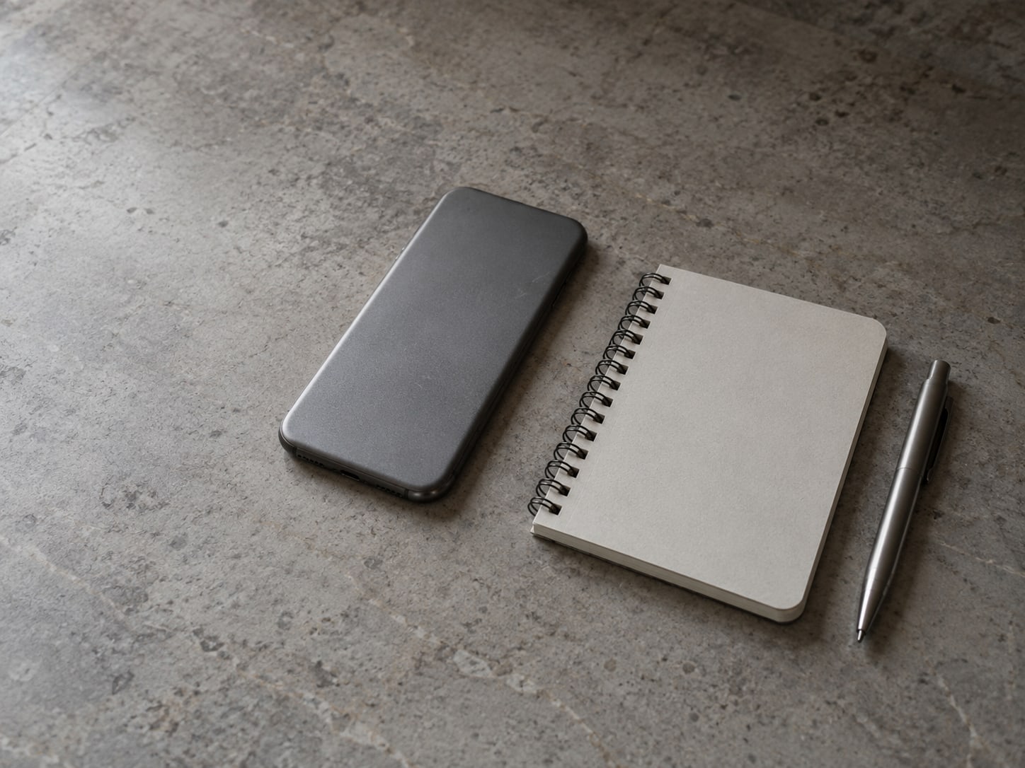 A closed phone lying face down beside a small spiral notebook and a ballpoint pen on a counter.