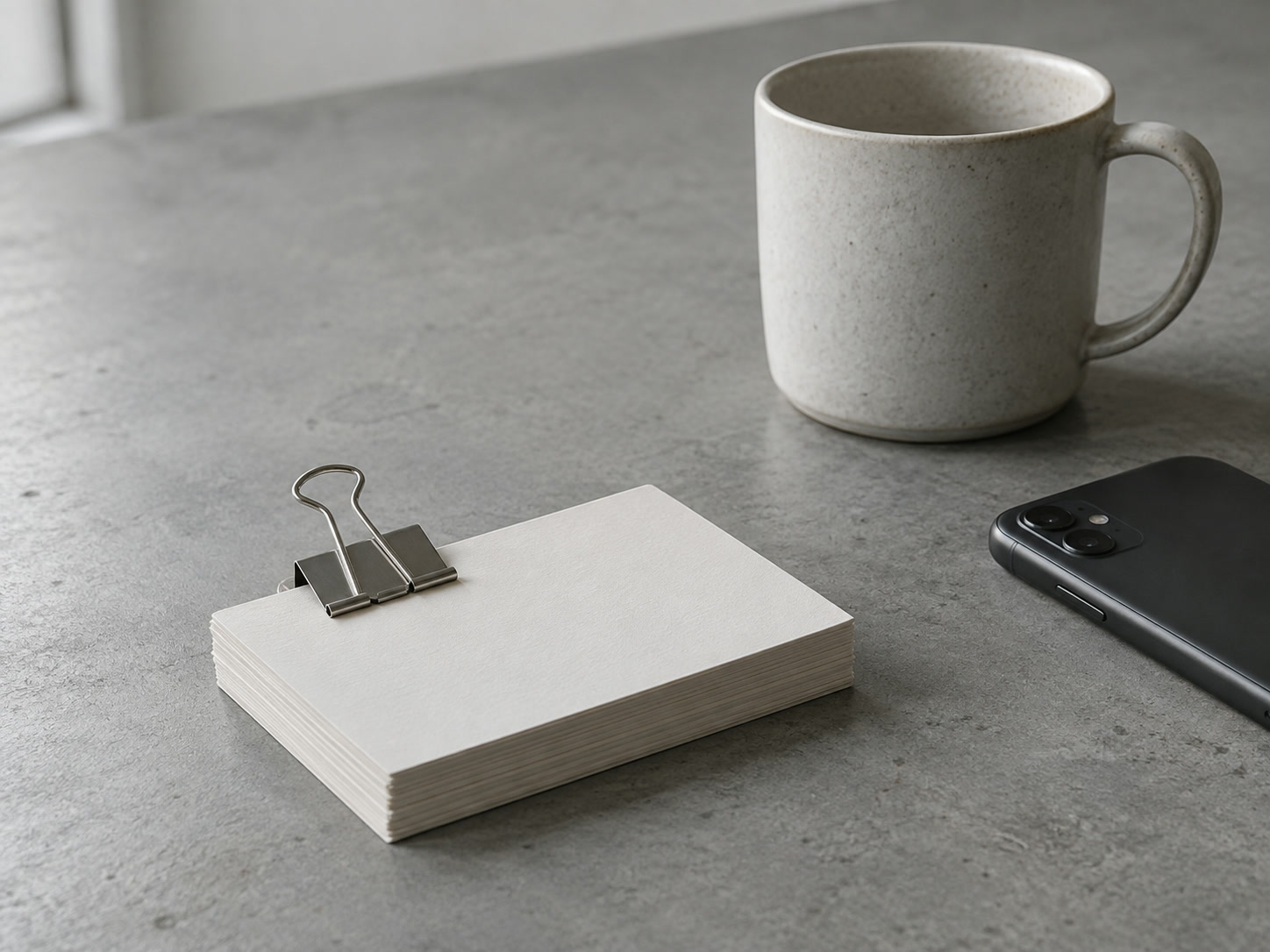 Una pila de tarjetas en blanco sostenidas con un clip junto a una taza y un teléfono boca abajo sobre una mesa gris.
