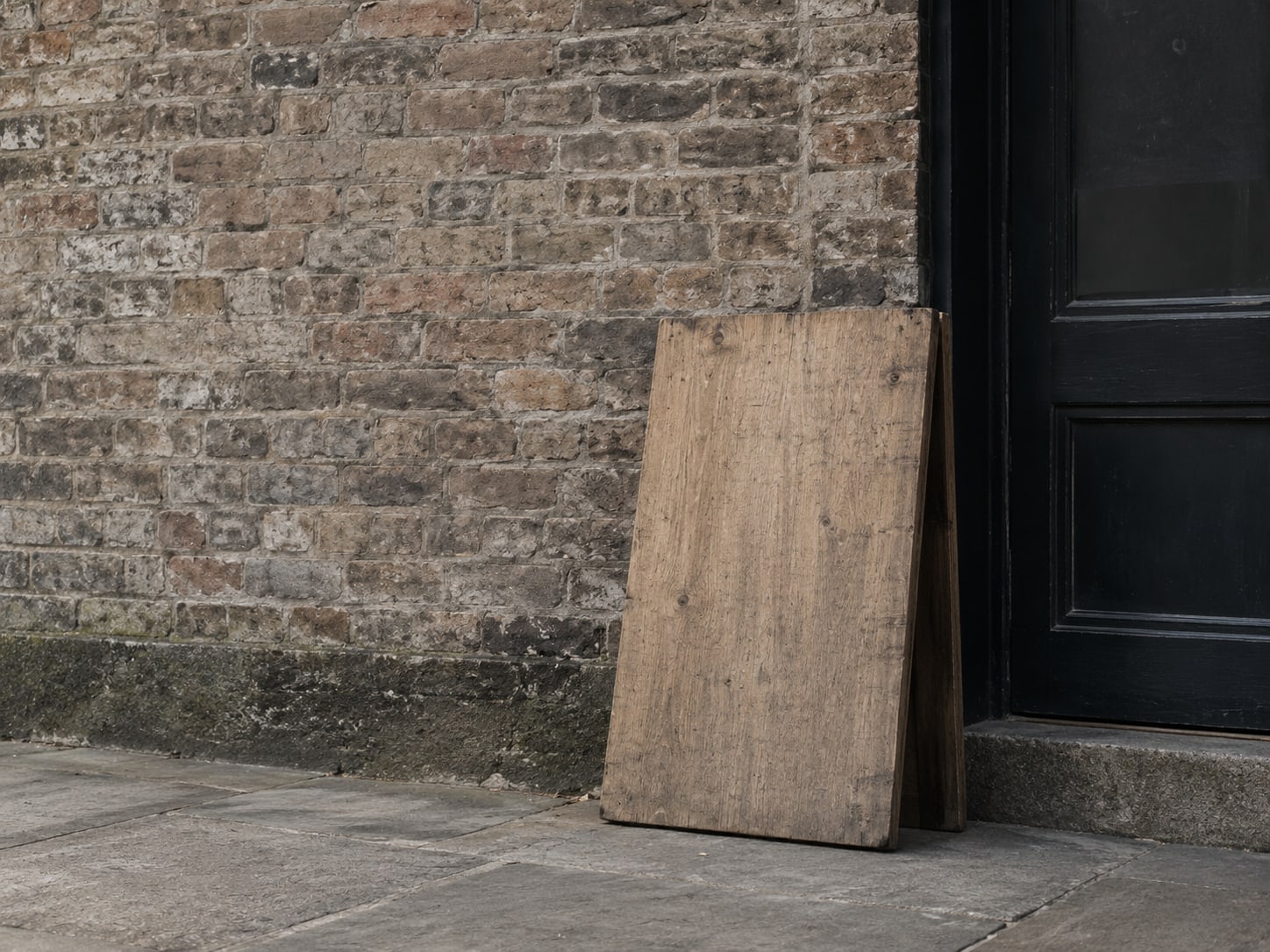 A blank wooden signboard leaning against a brick wall beside a closed shop door.
