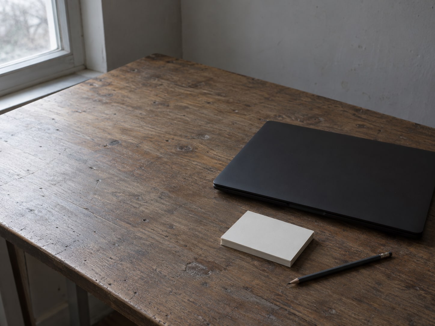A closed laptop beside a small stack of blank index cards and a pencil on a worn wooden desk.