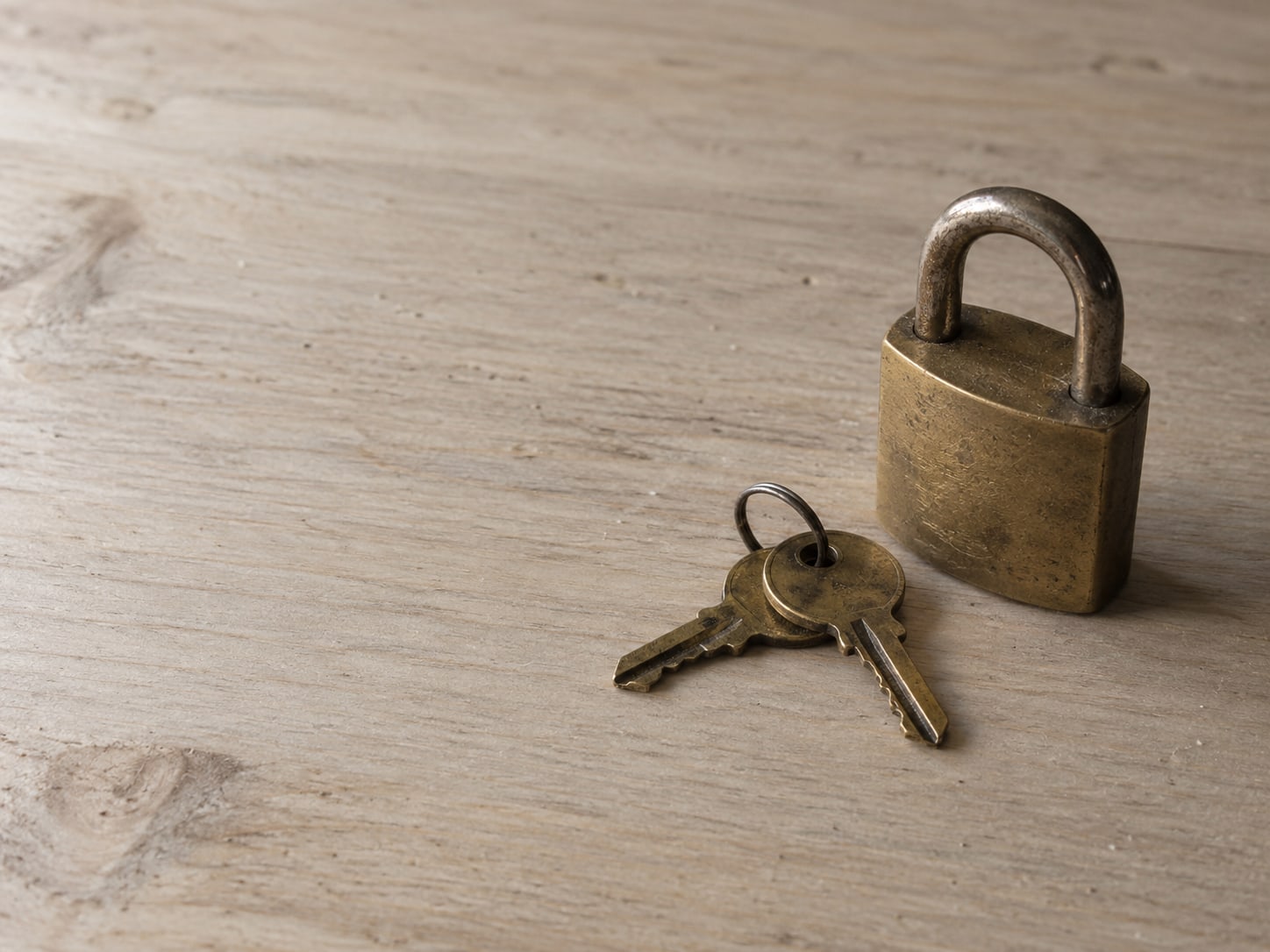 A pair of small brass keys on a plain wooden table beside a single closed padlock.