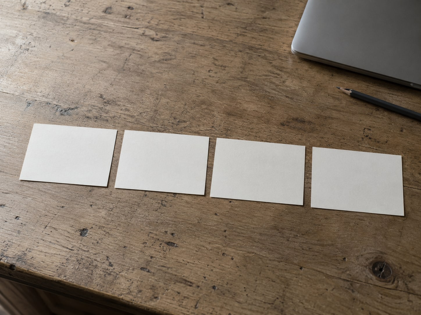 Four plain index cards laid in a row on a wooden desk beside a closed laptop and a pencil.