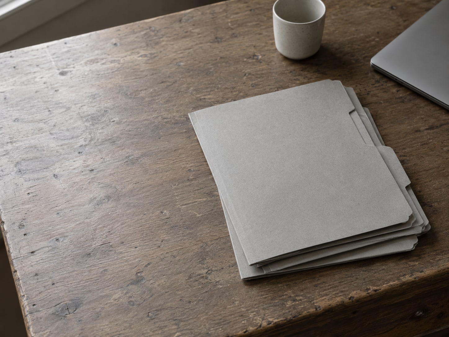 A tidy stack of blank folders beside a closed laptop and a ceramic cup on a wooden desk.