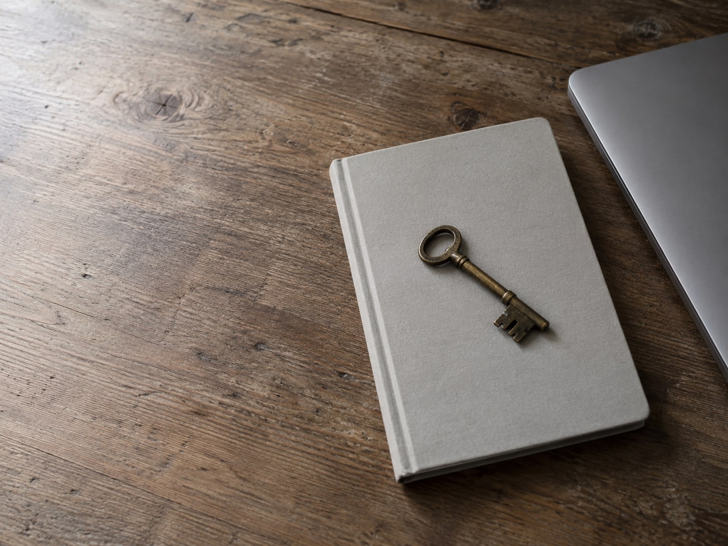 A small brass key resting on a blank notebook beside a closed laptop on a wooden table.