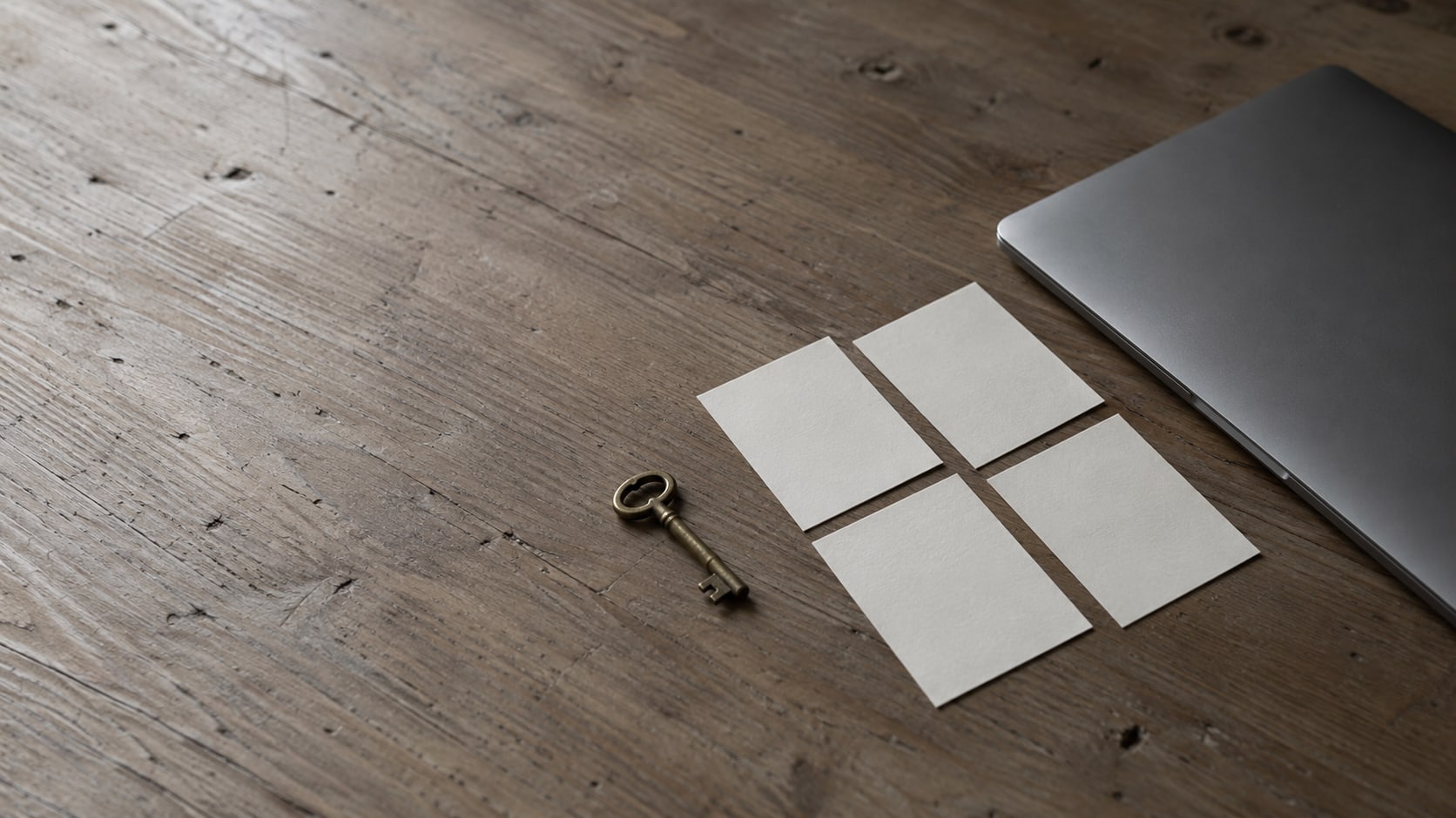 A closed laptop beside four blank index cards and a small brass key on a plain wooden desk.