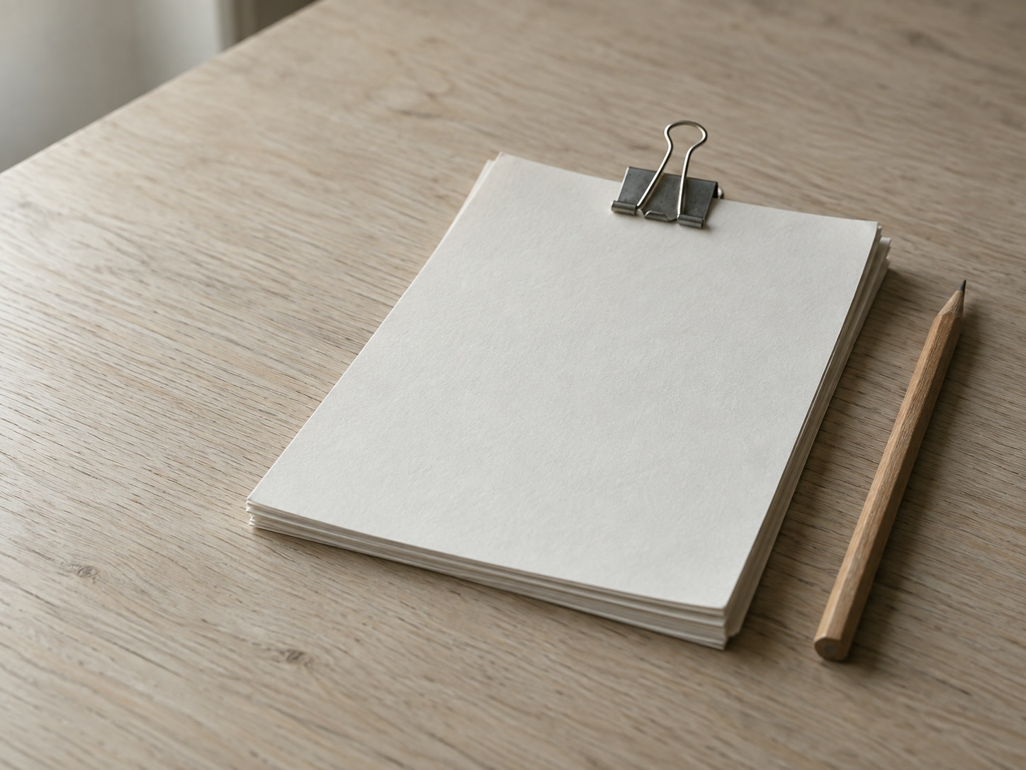 A short stack of blank paper forms held by a binder clip next to a sharpened pencil on a light oak table.