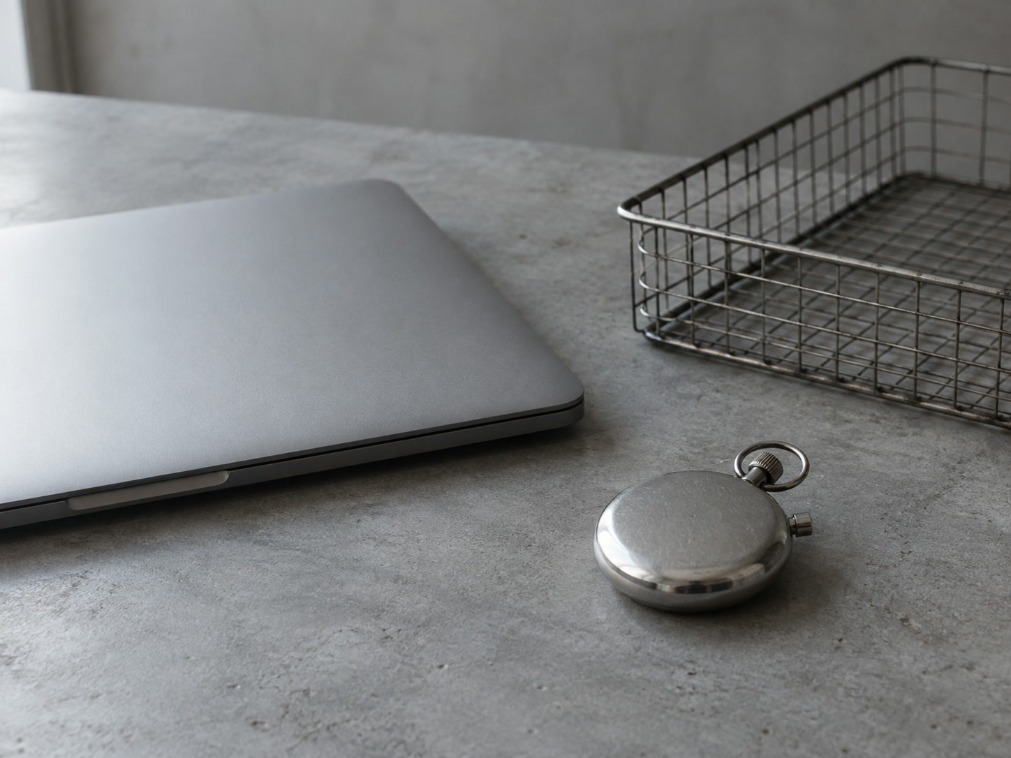 A closed laptop beside a stopwatch lying face down and an empty wire tray on a gray desk.