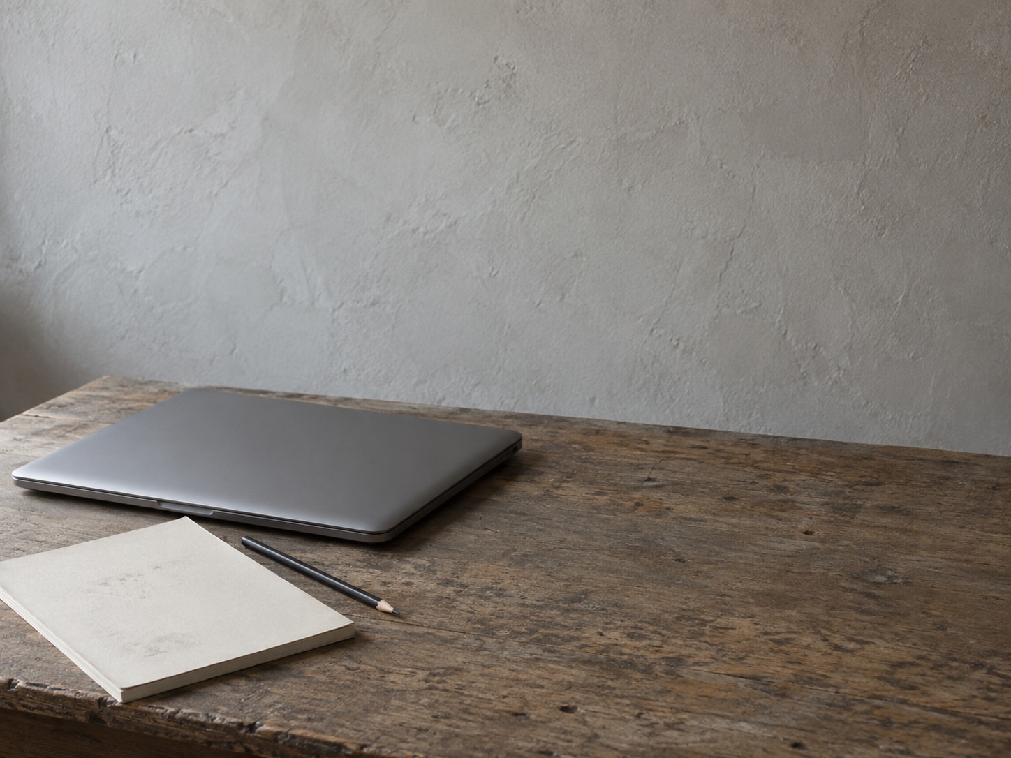 A closed laptop beside a blank notepad with faint pencil marks and a pencil on a worn wooden desk.