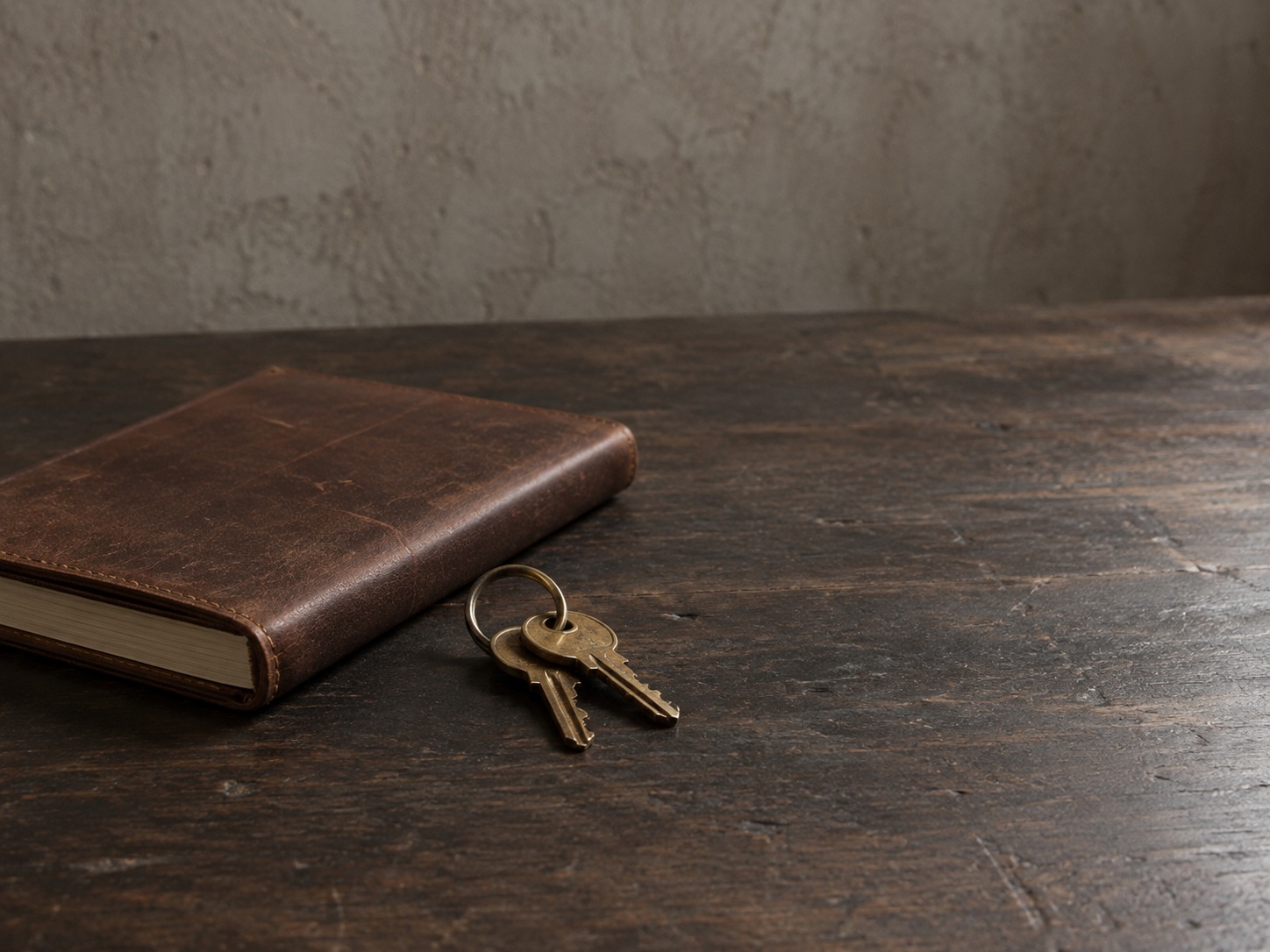 Two small brass keys on a plain ring resting beside a closed leather notebook on a dark wooden desk.
