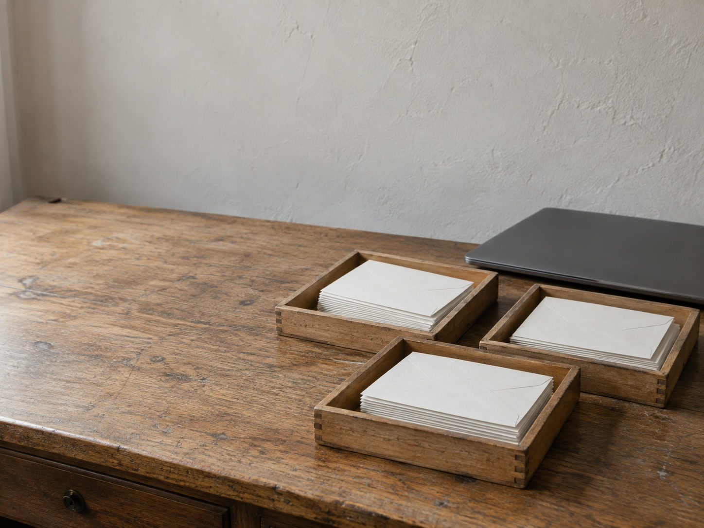 Three shallow wooden trays on an oak desk, each holding a small stack of blank envelopes, beside a closed laptop.