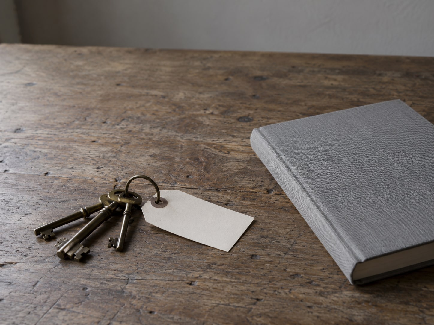 A small ring of brass keys beside a blank paper tag on a wooden desk, with a closed notebook next to them.