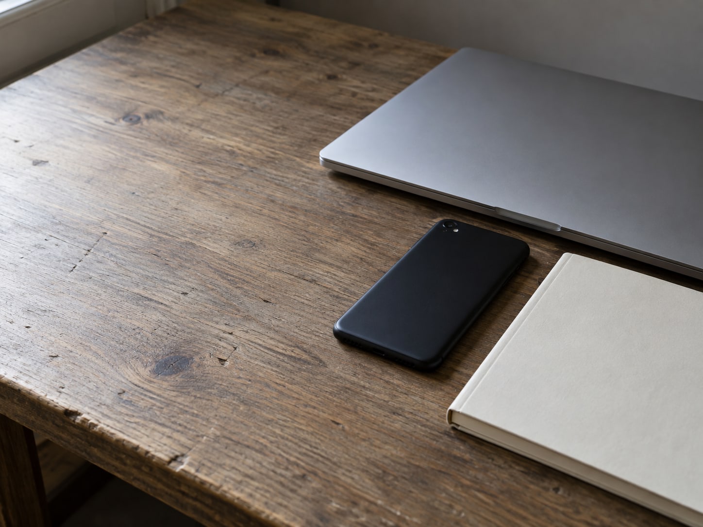 A closed laptop and a switched-off Android phone resting face down beside a plain notebook on a wooden desk.