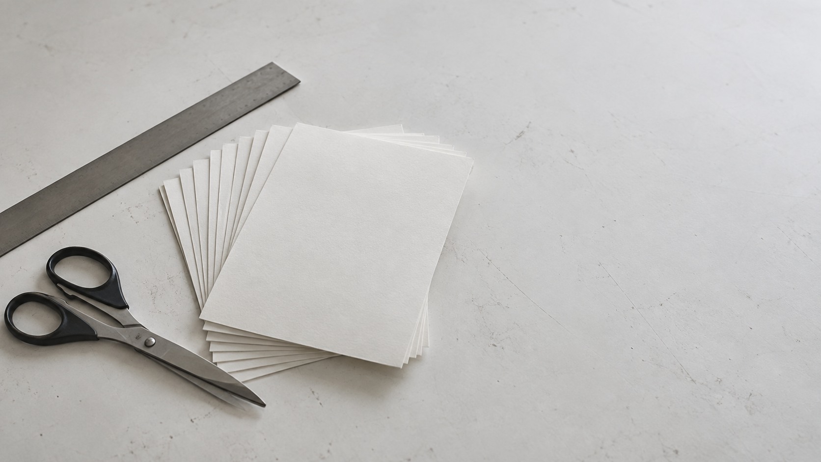 A fanned stack of blank printed card stock beside a pair of scissors and a metal ruler on a white table.