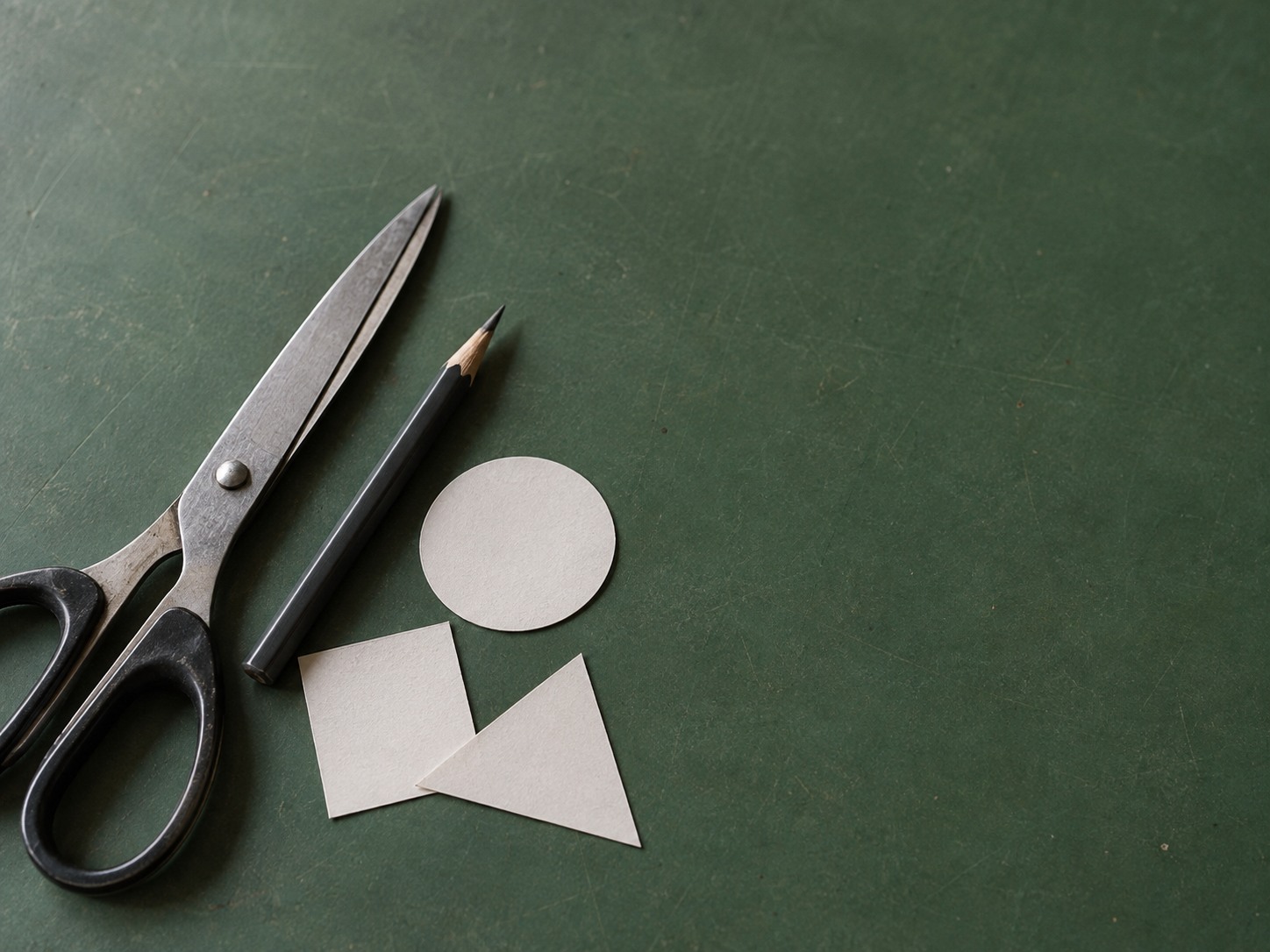 Unas tijeras, un lápiz afilado y unos cuantos recortes de papel en blanco dispuestos sobre un tapete de corte verde.