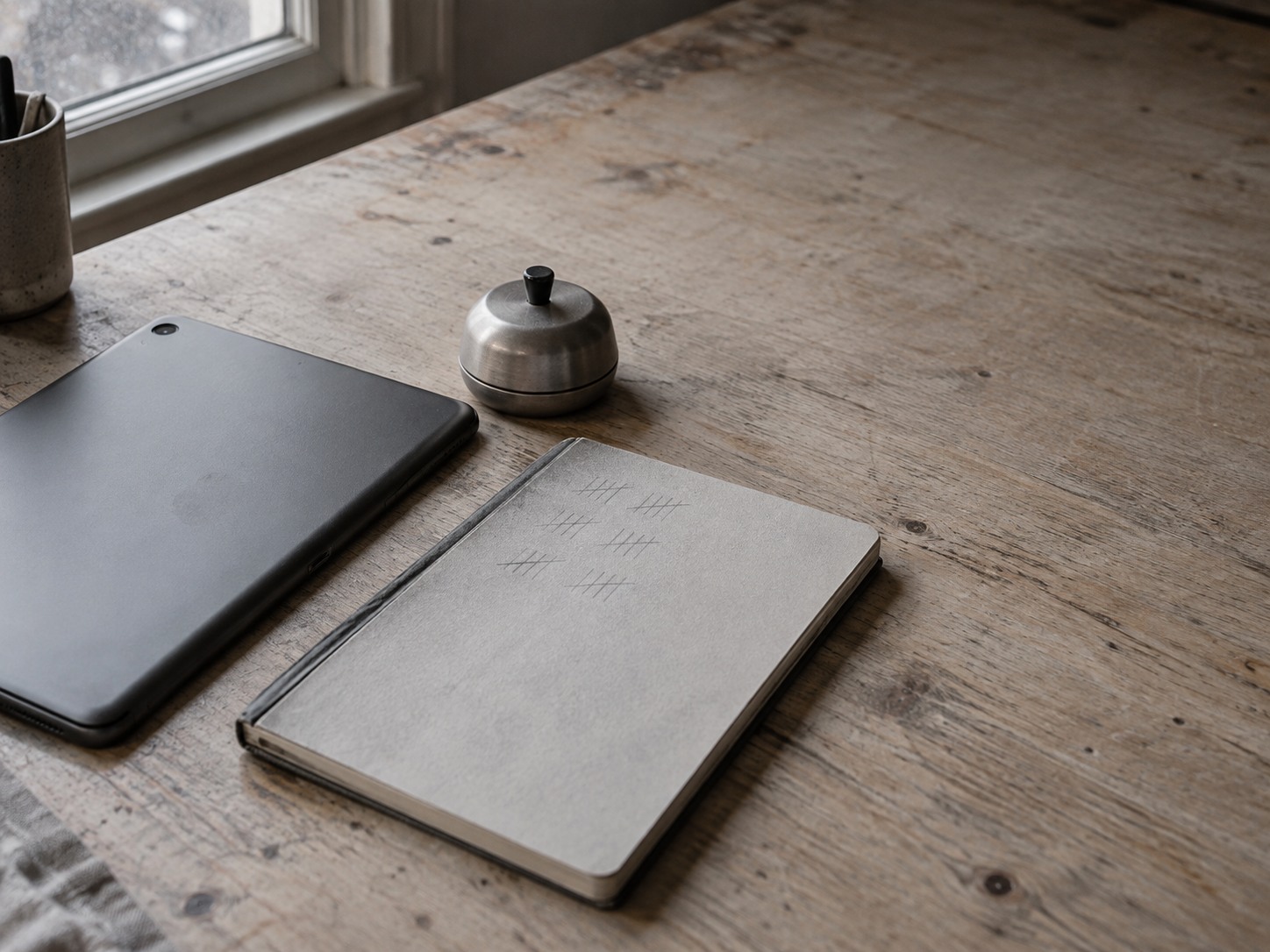 Una libreta con marcas de lápiz tenues junto a una tableta boca abajo y un pequeño temporizador de cocina sobre un escritorio de madera.