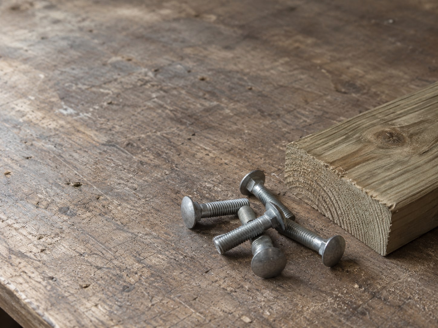 A small pile of galvanized carriage bolts beside a short length of pressure-treated lumber on a workbench.