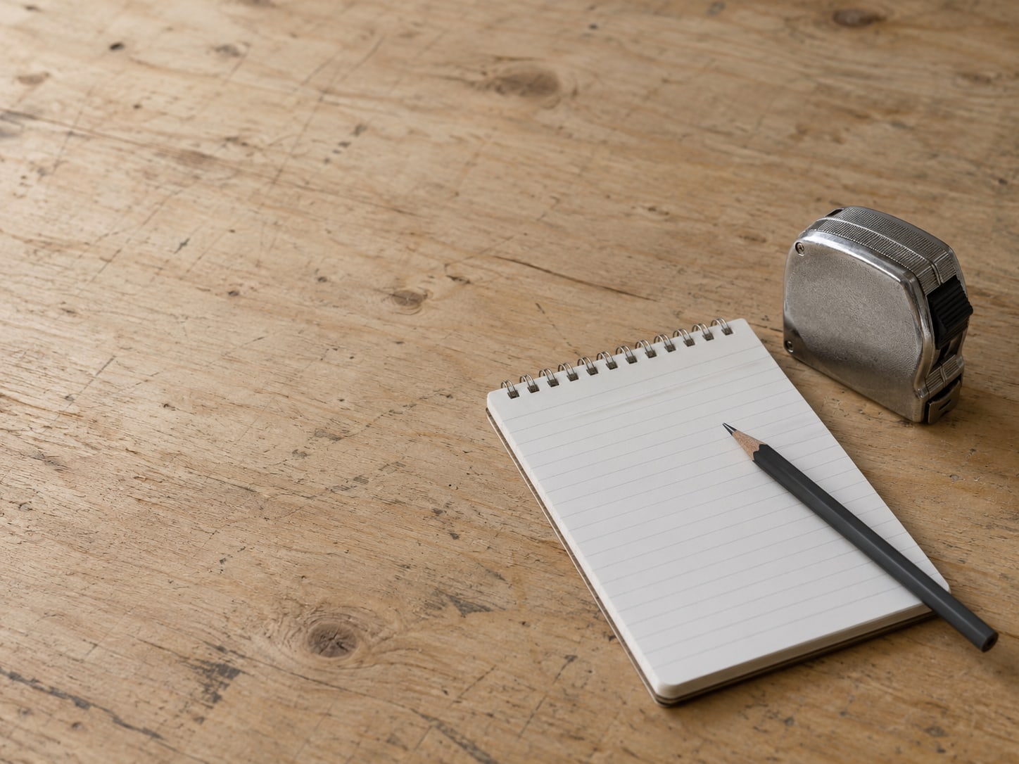 A closed steel tape measure and a pencil resting on a blank ruled notepad on a plywood tabletop.