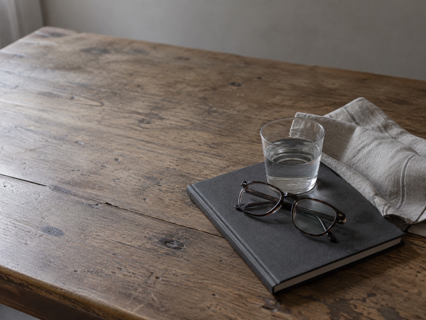 Una servilleta de lino doblada, un vaso pequeño de agua y unos lentes de lectura sobre un cuaderno cerrado, en una mesa de cocina bajo luz suave de la mañana.