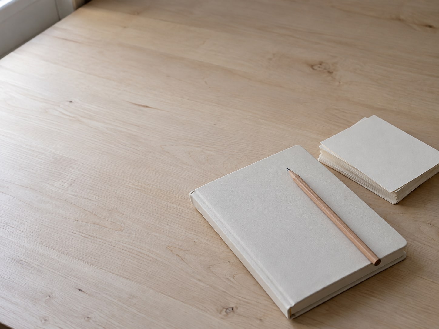 A closed notebook with a pencil resting on top, next to a small stack of blank index cards on a pale wooden desk.