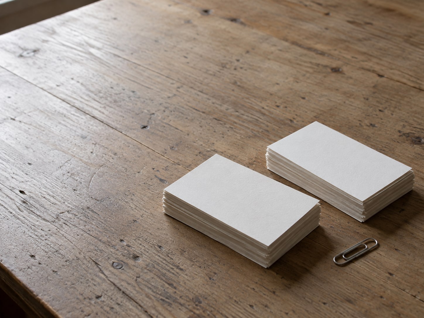Two stacks of blank index cards sorted into separate piles on a worn wooden desk, with a small paper clip beside them.