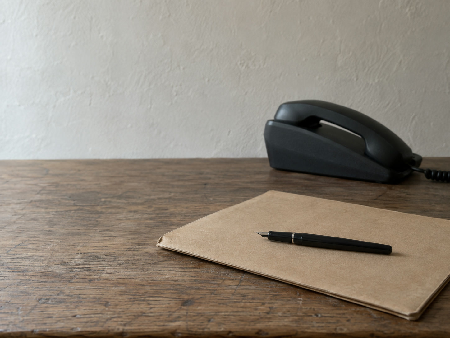A closed paper folder and a fountain pen resting on a desk beside a desk phone with its handset down.