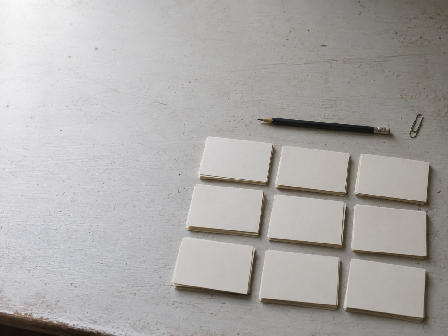 A pencil, a paper clip and a small stack of unmarked index cards laid out in three rows on a pale desk.