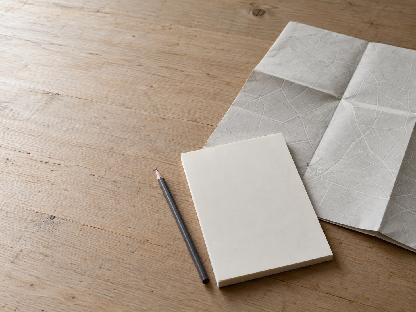 Un lápiz, una libreta en blanco y un mapa de carreteras de papel doblado sobre una mesa de madera lisa; el mapa está abierto en una zona de calles sin marcar.