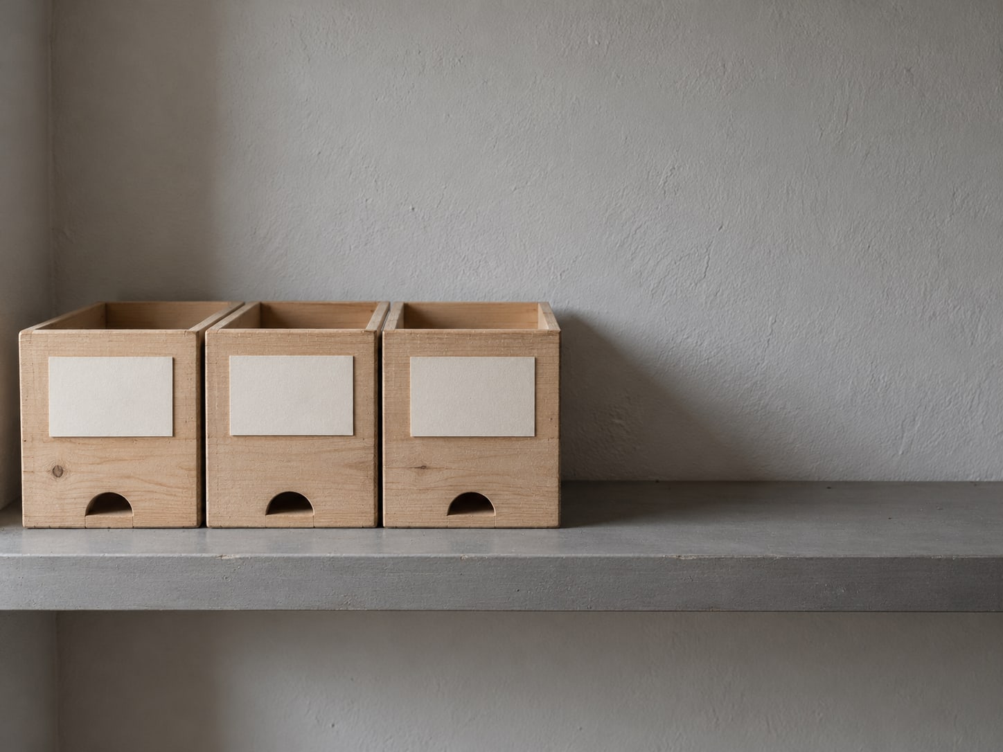 A tidy row of three wooden filing boxes with blank paper labels on a bare shelf.