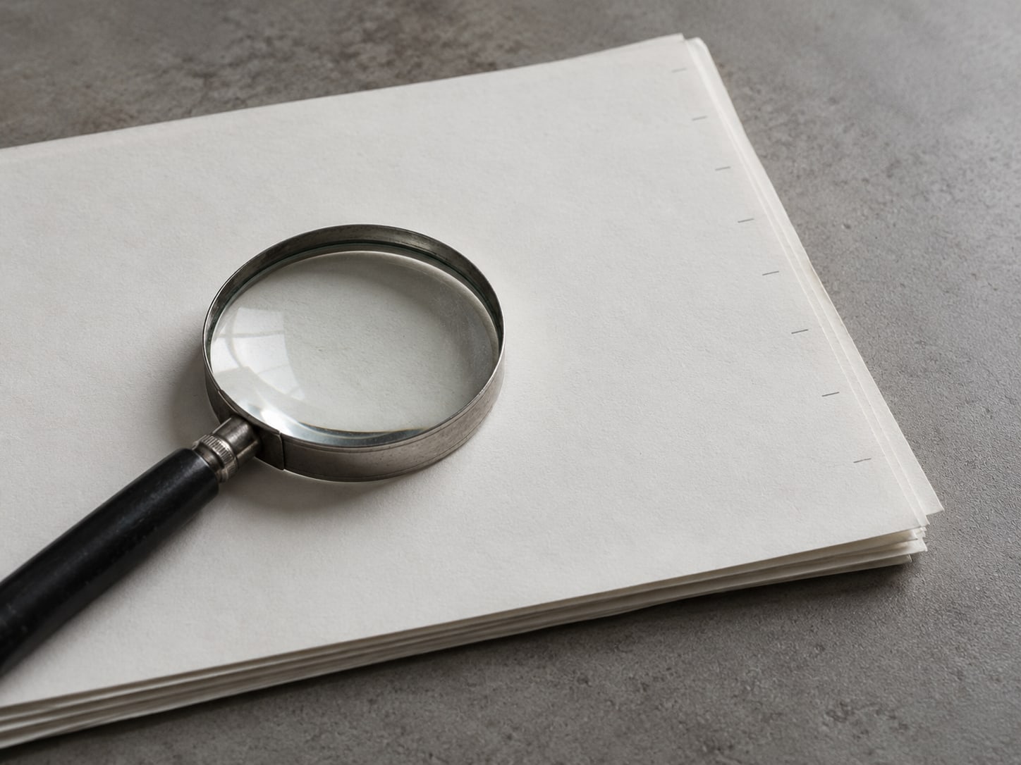A magnifying glass resting on a stack of blank paper with faint pencil ticks along one edge.