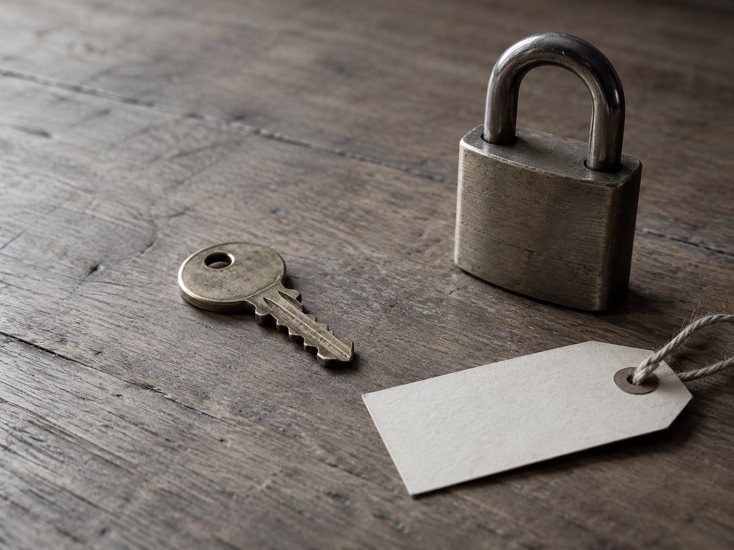 A single brass key on a wooden desk beside a closed padlock and a blank paper tag.