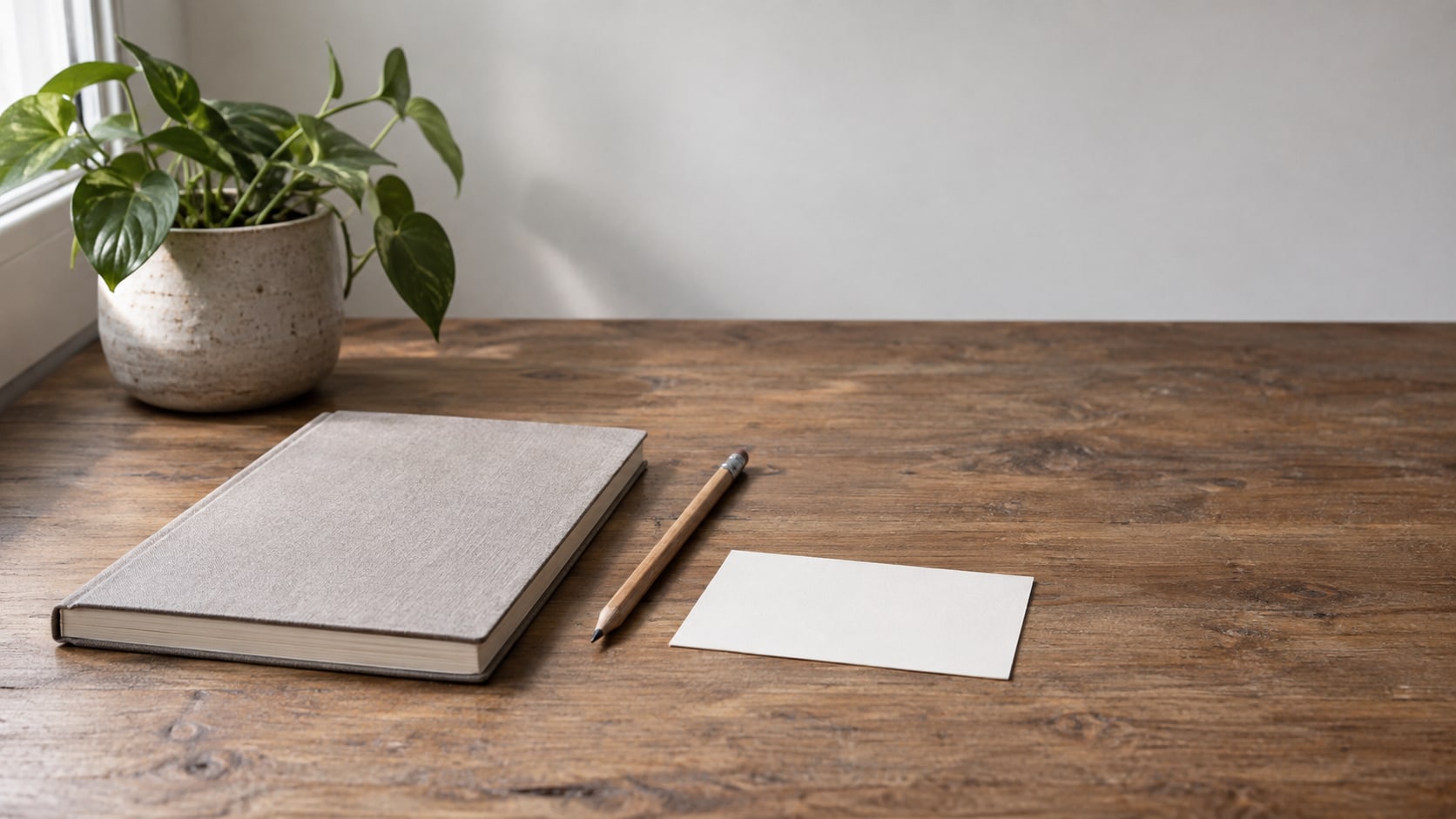 A closed notebook, a pencil and a small potted plant on a wooden desk beside a blank index card.