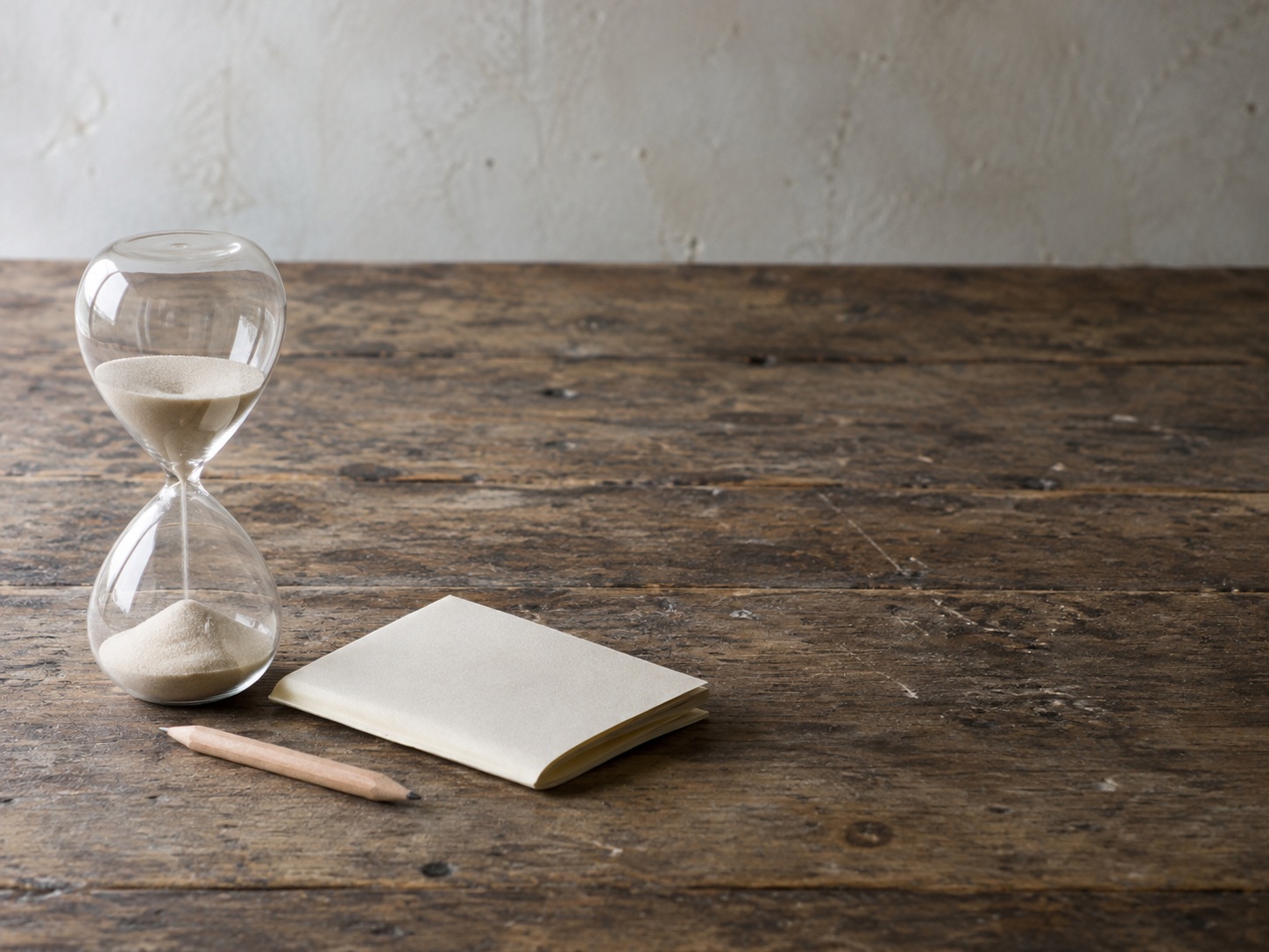 An hourglass beside a folded blank sheet of paper and a short pencil on a worn wooden workbench.
