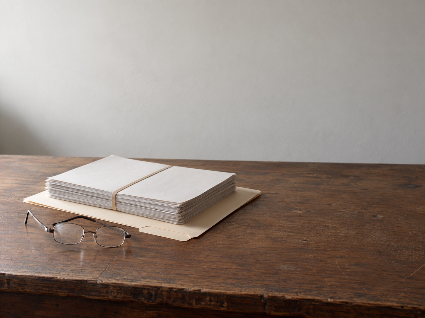 A closed manila folder, a rubber-banded stack of blank papers and a pair of reading glasses on a plain wooden desk.
