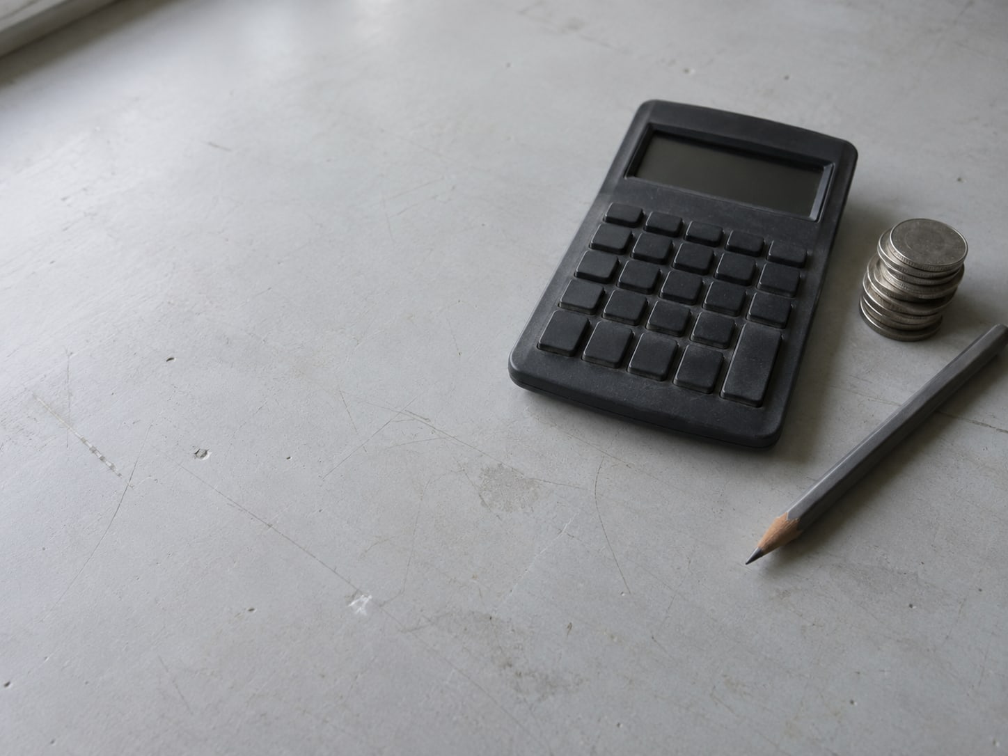Una calculadora apagada junto a una pequeña pila de monedas y un lápiz sobre un escritorio liso.