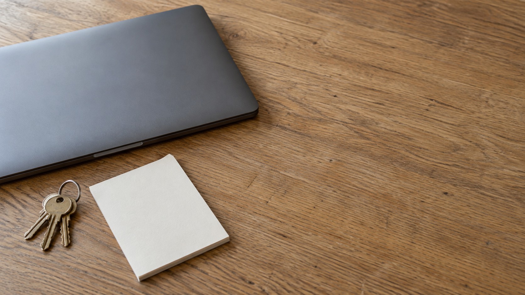 A closed laptop beside a small ring of keys and a blank notepad on a clean wooden desk.