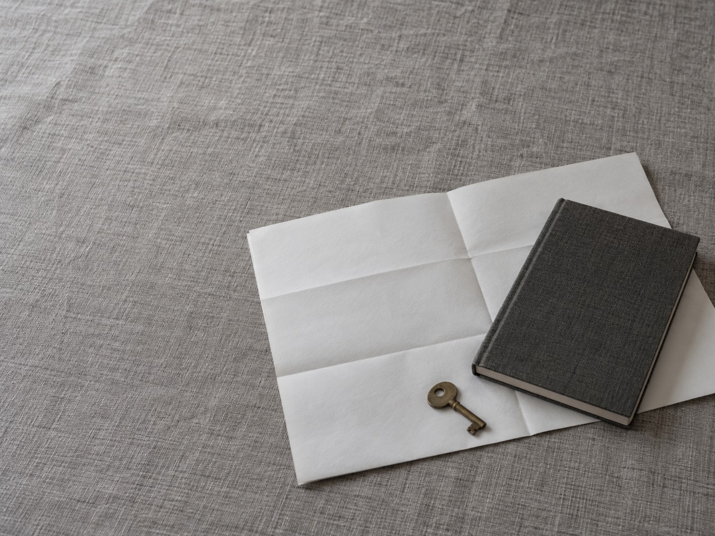 A closed notebook resting on a folded paper floor plan beside a small brass key on a gray linen tablecloth.