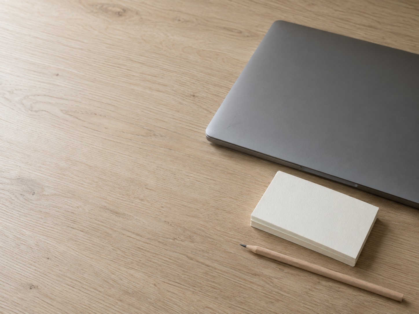 A closed laptop beside a short stack of blank index cards and a pencil on a pale oak desk.