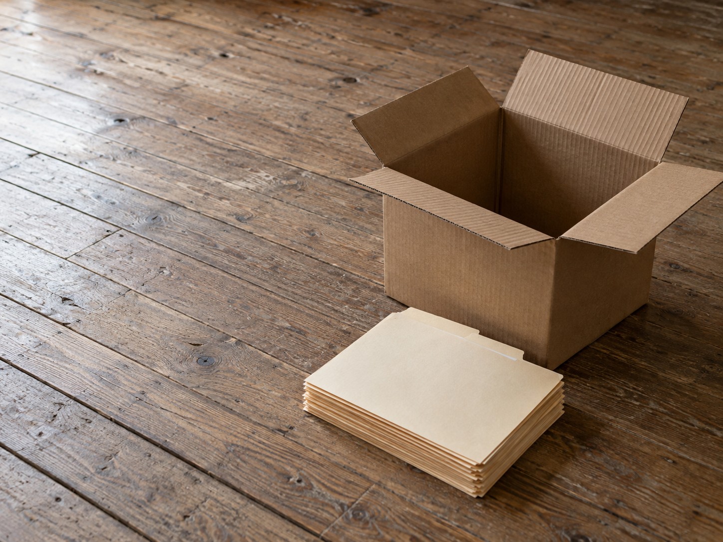 A cardboard moving box with its flaps open beside a neat stack of blank manila folders on a wooden floor.