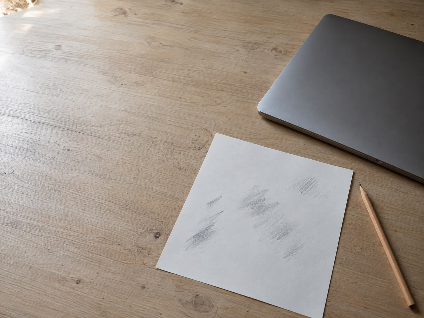 A blank sheet of paper with faint pencil marks beside a sharpened pencil and a closed laptop on a wooden desk.