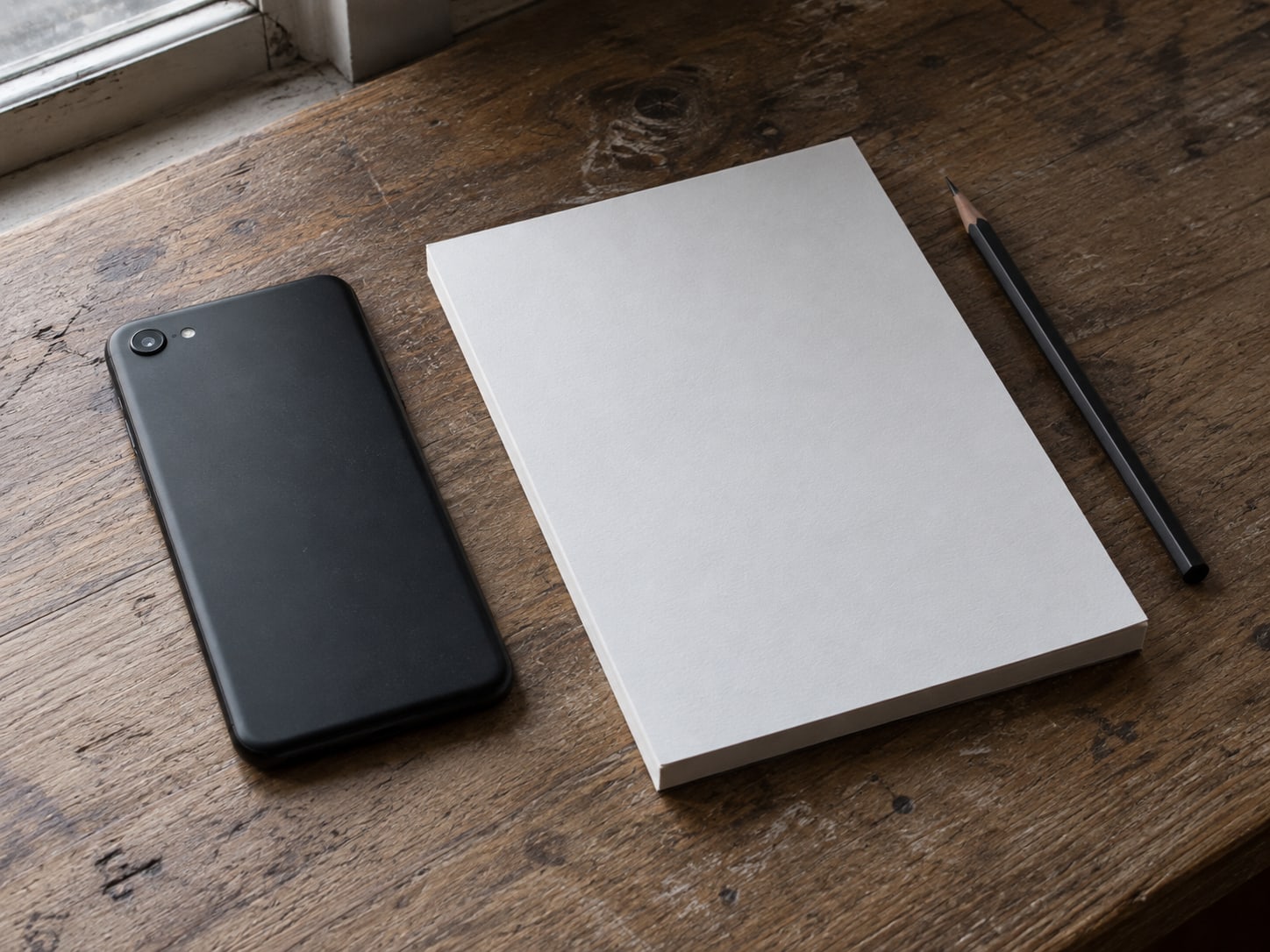 A phone lying face down on a wooden desk beside a blank notepad and a pencil.