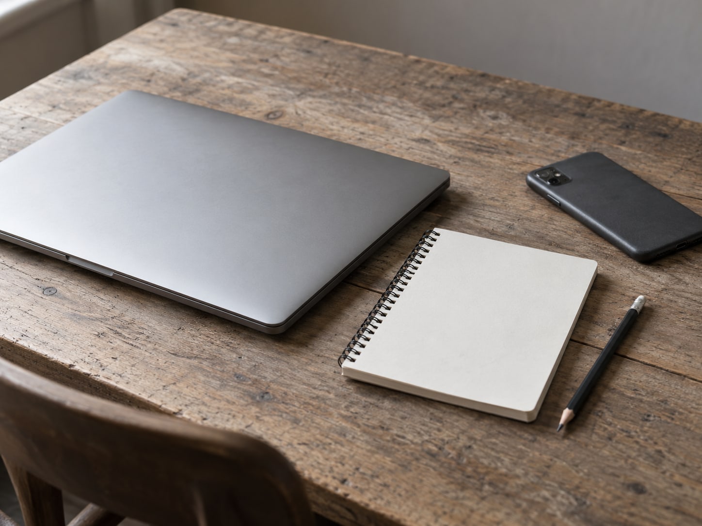 A closed laptop beside a small spiral notebook and a pencil on a wooden desk, with a phone lying face down next to them.