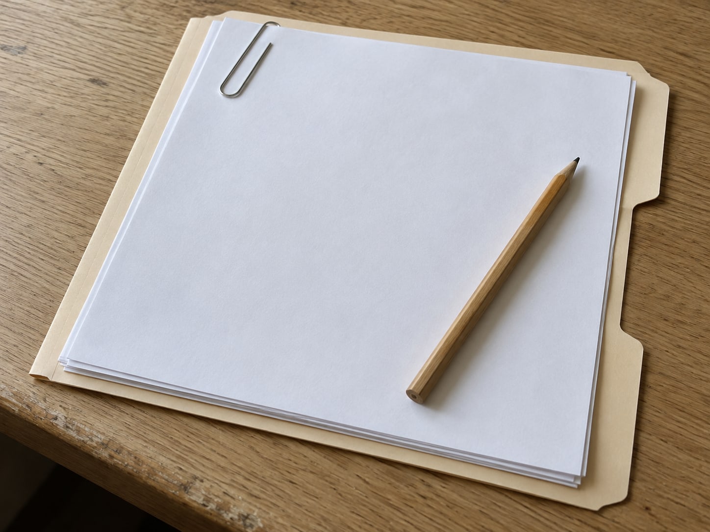 A manila folder holding a neat stack of blank printed pages, with a paper clip and a pencil resting on top, on a light oak table.