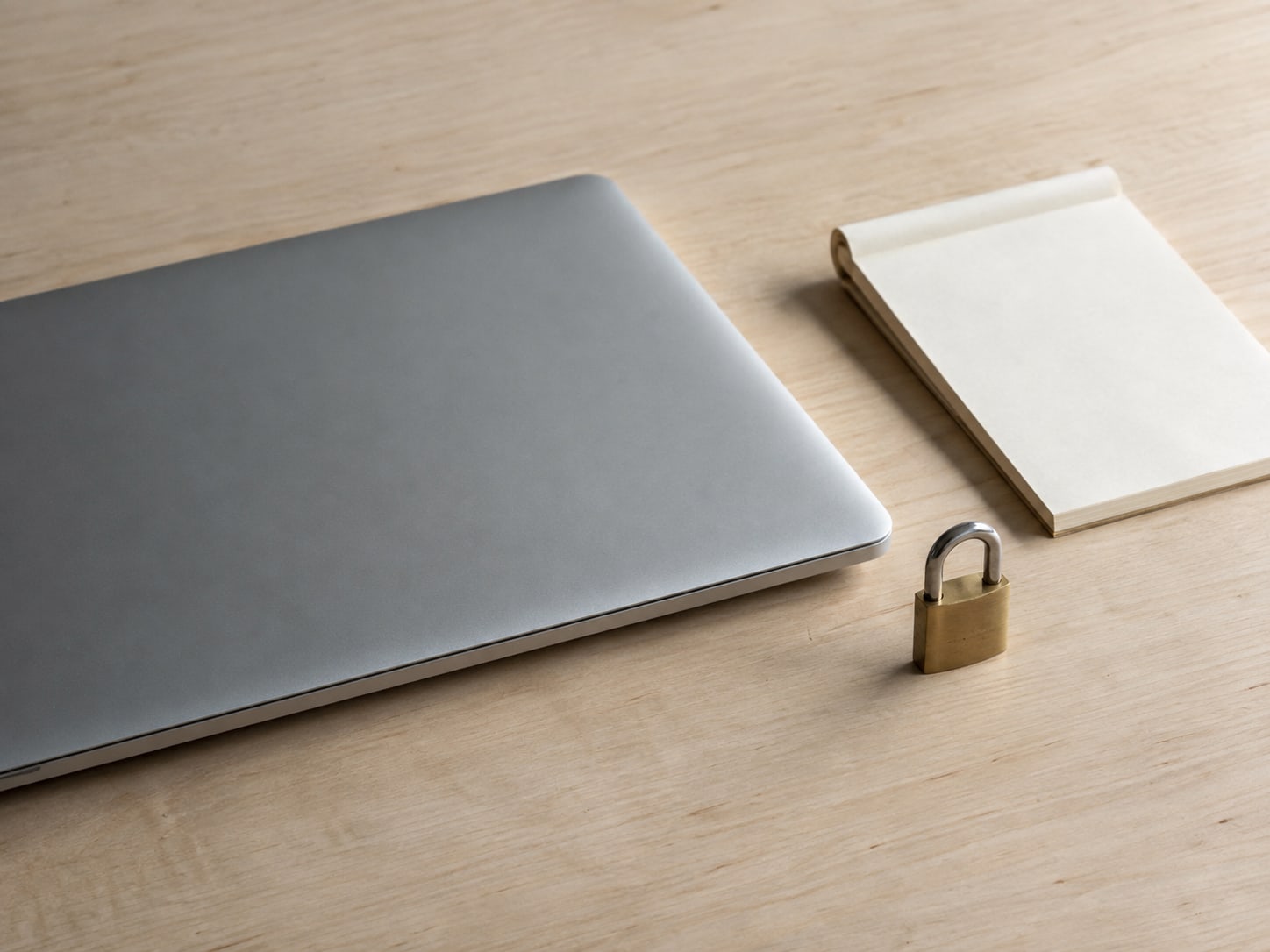 A closed laptop on a clean wooden desk beside a small brass padlock and a folded paper notepad with blank pages.
