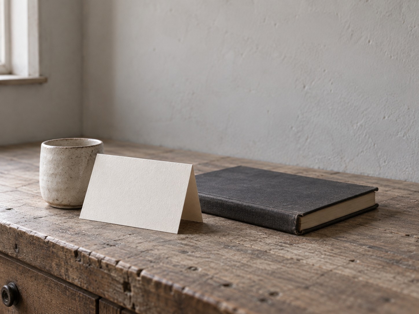 A folded blank notice card propped against a small ceramic cup on a shop counter beside a closed ledger.