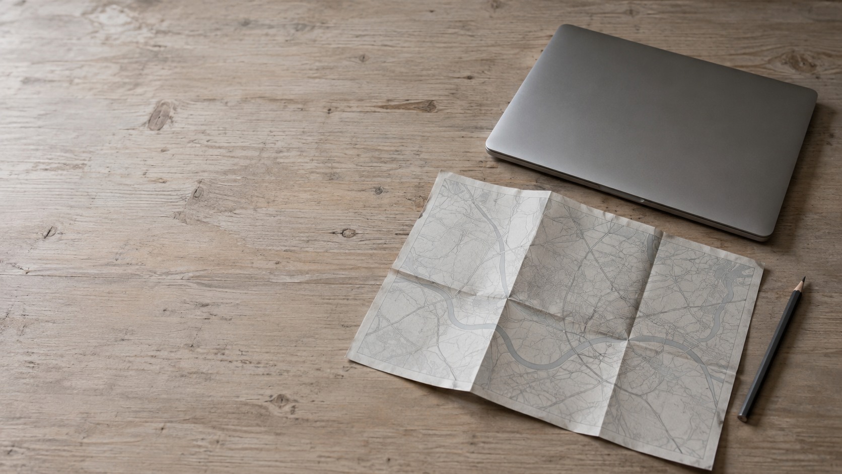 A folded paper map with no pins in it beside a closed laptop and a pencil on a light wooden desk.