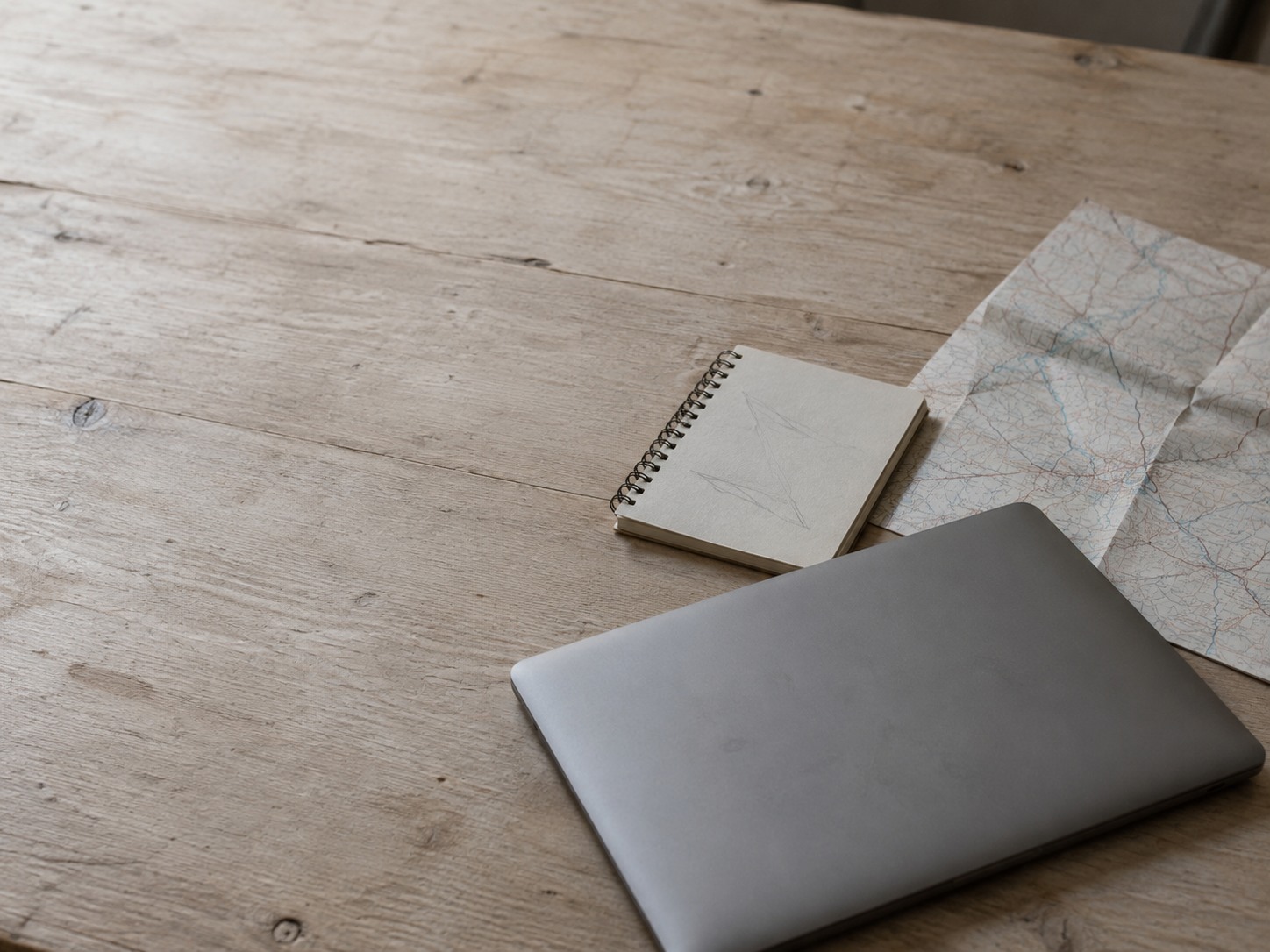 A closed laptop and a small spiral notebook with faint pencil marks resting beside a folded paper street map on a light wooden table.