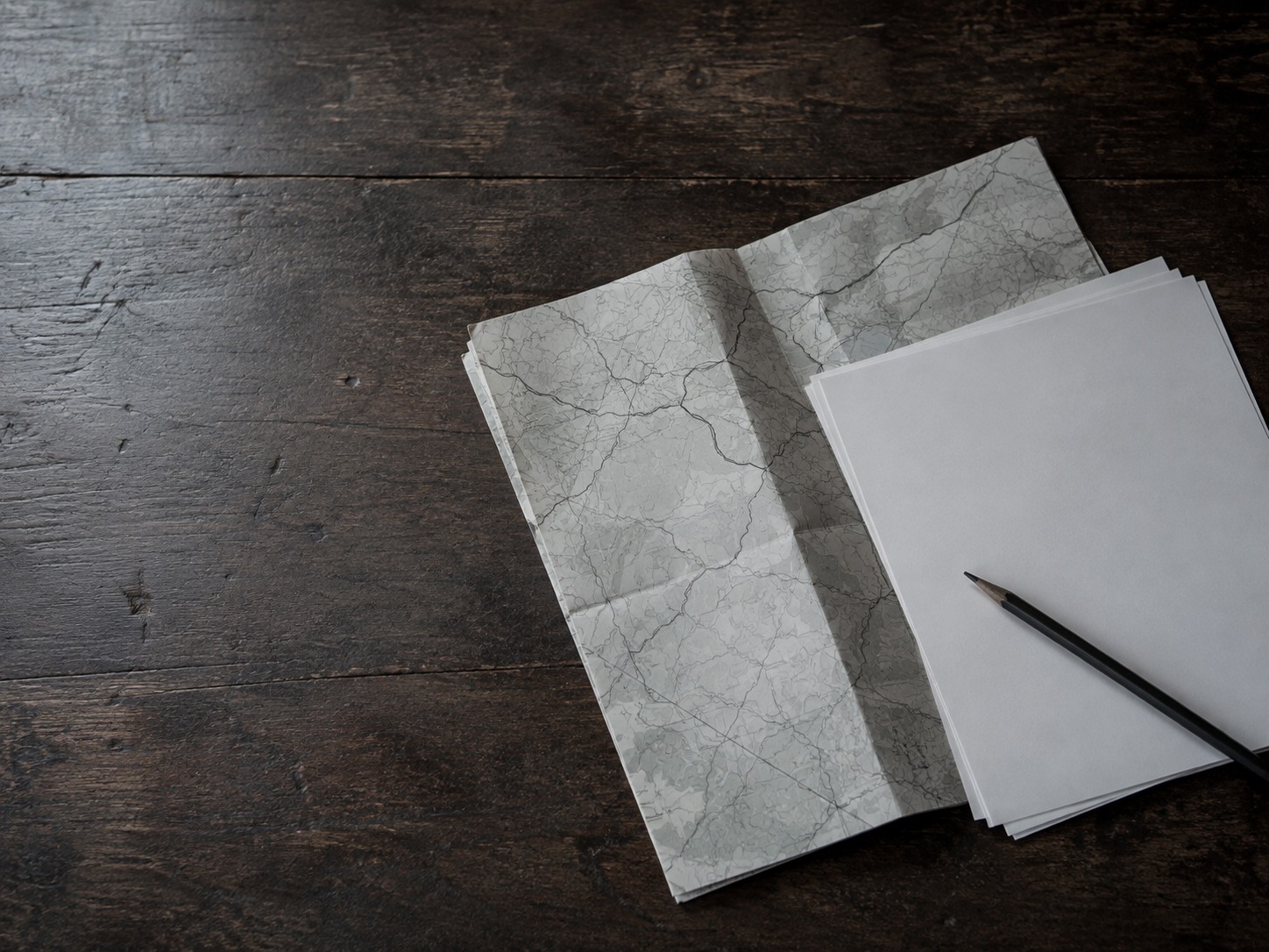 A single folded paper map lying beside a thin stack of blank survey sheets and a pencil on a dark wooden desk.